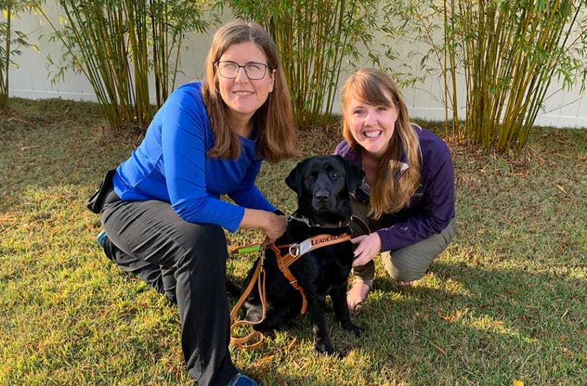 Two women kneel outdoors with a black lab in Leader Dog harness between them. They are both smiling. The left woman is wearing glasses. The woman on the right is instructor Ashley