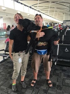 Ashley stands in an airport seating area with a man standing next to her. He is holding up a black lab in Leader Dog harness. The dog is giving Ashley a kiss.