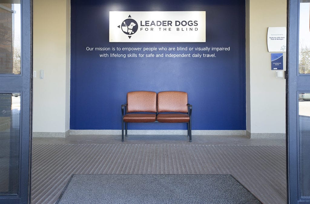 Photo shows the entrance to the canine center on Leader Dog's campus. There is a blue wall with Leader Dog's logo on a white plaque. Beneath in white lettering is our mission statement. There is a brown double chair on the floor.