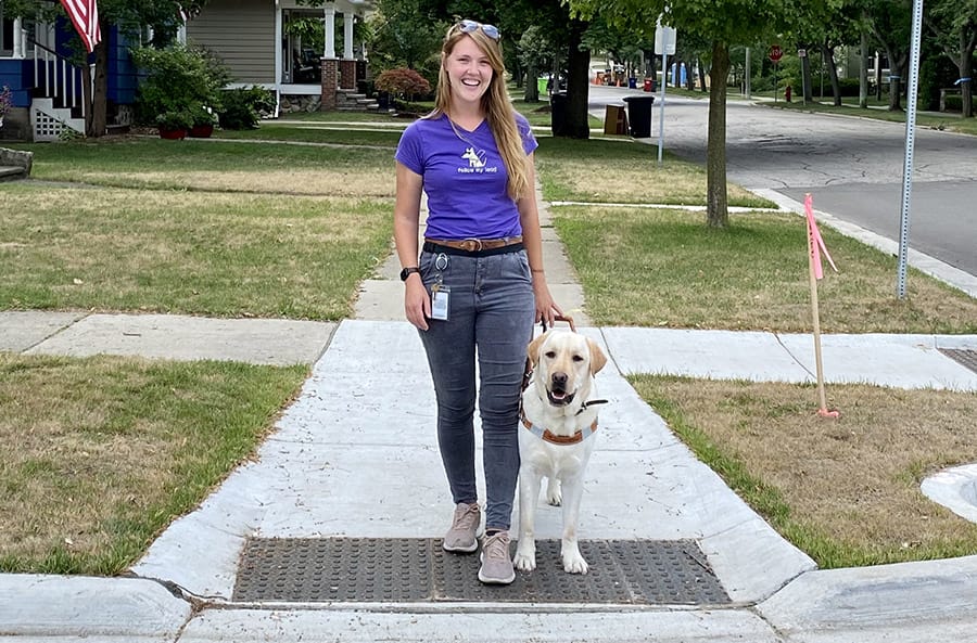 Ashley is standing at a curb on the sidewalk of a tree-lined street. She is smiling at the camera and walking with a yellow lab in Leader Dog harness.