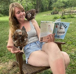 Ashley sits on a chair in a grassy yard. She is holding a chicken in one arm and a book in the other hand titled "How to Speak Chicken." Another chicken is perched on her shoulder.