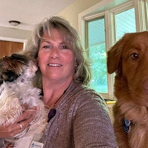 Photo of Barrie Lynn smiling at the camera. She is holding a small dog and there's a large, reddish dog next to her that appears to be a golden retriever.