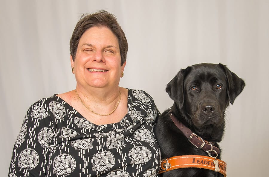 Joanne sits in front of a gray photo background. She is wearing a black and white flowery shirt and smiling. Next to her is a seated black lab in harness.
