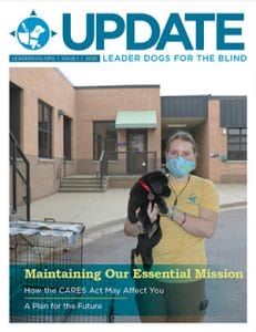 Cover of Update issue 1 - July 2020. Photo shows a young woman wearing a light blue mask and a yellow t-shirt with the Leader Dog logo holding a black lab puppy outside a brick building.