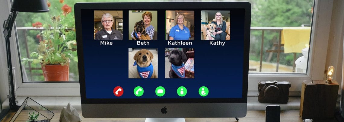 Photo of computer monitor on a desk. On the monitor is a Zoom call with four people and two puppies pictured on the Zoom call.