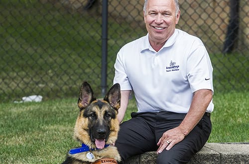 Rod sits on a low wall outside near green grass. He is wearing a white polo with a vintage Leader Dog logo and smiling at the camera. A German shepherd in Leader Dog harness sits next to Rod on the ground.