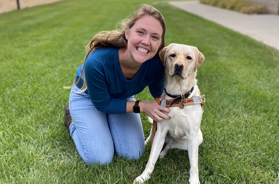Ashley kneels on a lawn, smiling, with her arms around a yellow lab wearing a Leader Dog harness. The dog is sitting next to her and looking at the camera