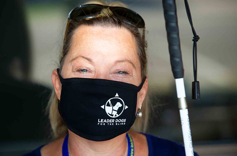 Close-up photo of a woman looking into the camera wearing a black face mask with a white Leader Dog logo on the left side. There are crinkles around her eyes that make it appear she may be smiling. She is holding a white cane.