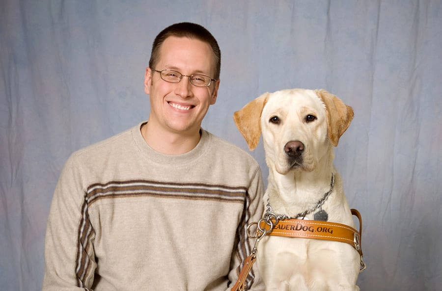Andy sits smiling in front of a gray photo backdrop with yellow lab Ingrid sitting next to him. Ingrid is in her Leader Dog harness and Andy holds her leash.