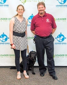 Phil stands next to a female client and her black lab Leader Dog in front of a photo background with repeating blue and green Leader Dog logos.