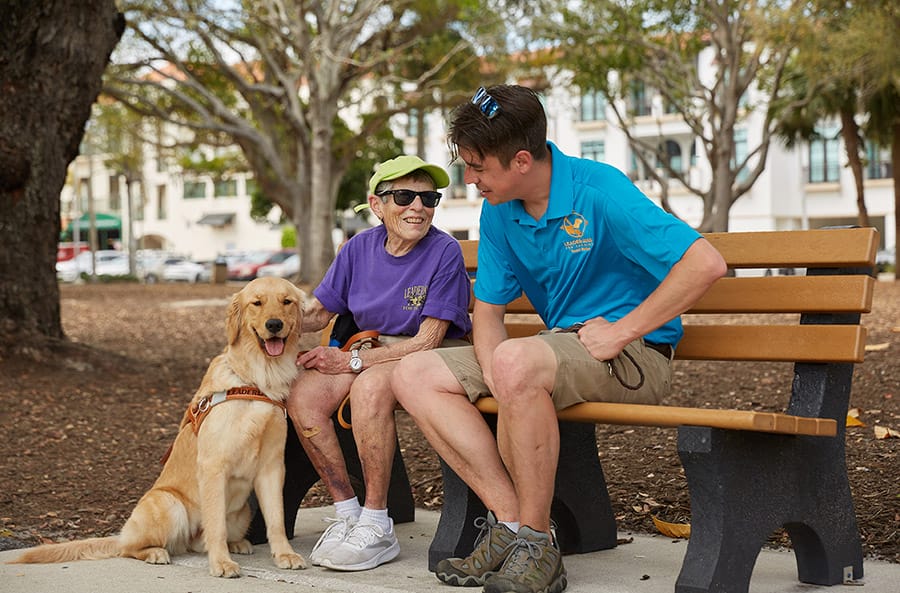 A male Leader Dog guide dog mobility instructor sits on a bench with a female client. They are smiling and appear to be talking. A golden retriever in leather Leader Dog harness sits next to the client on the ground.