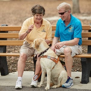 Keith sits on a bench with a female client. They are smiling and looking down toward a yellow lab in Leader Dog harness that is sitting on the sidewalk in front of them.