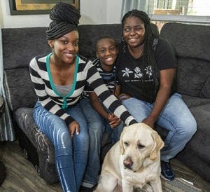 Leatrice and her son and daughter sit on a gray couch in a living room. All are smiling at the camera. Leatrice's daughter has her hand on a yellow lab that is sitting on the floor in front of them.