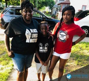 Leatrice stands on a sidewalk with her son and daughter. All three of them are smiling at the camera. Leatrice is wearing a Black Lives Matter t-shirt and her son and daughter are wearing t-shirts in celebration of Juneteenth.