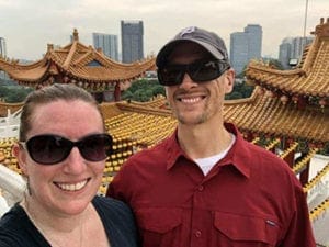 Andy and his wife stand in front of building with a decorative roof in Malaysia. They are both smiling at the camera.