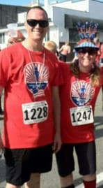 Andy and a female friend stand next to each other in matching red shirts. They are smiling and both wearing race numbers on the front of their shirts.