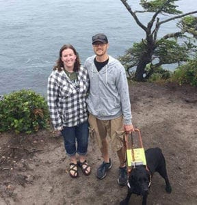 Andy, his wife and Leader Dog Rayna pose at the edge of a hill overlooking a large body of water.