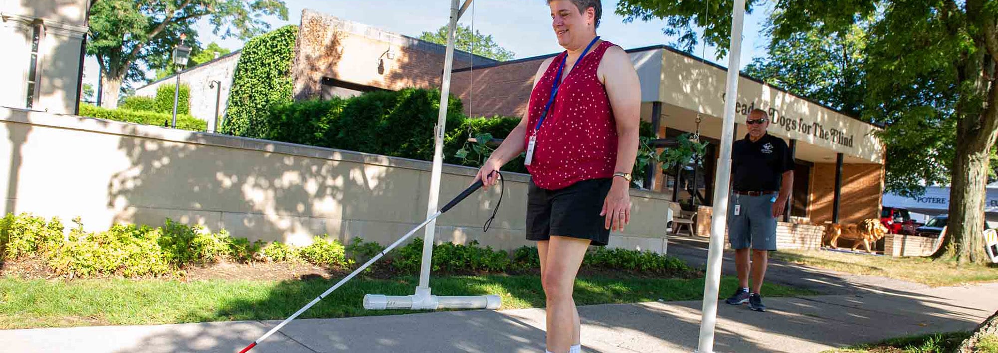 A woman walks on a grass and tree lined sidewalk on a sunny day. She is smiling and navigating with a white cane.