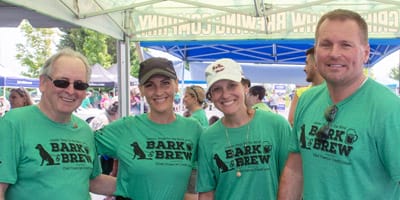 A group of two men and two women stand near each other, all smiling at the camera and wearing green t-shirts with Leader Dog's Bark & Brew event logo on the front