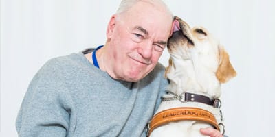 A older man sits in front of a white background, smiling at the camera. His face is being licked by a yellow lab in Leader Dog harness seated next to him.