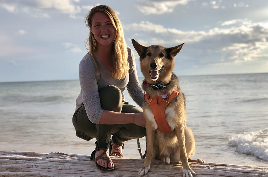Ashley kneels on a log in front of a large body of water in the background. Her hands are on a dog in an orange body harness next to her.
