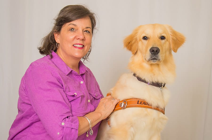 Audrey sits in front of a light gray photo backdrop. She is smiling at the camera and wearing a pink button up shirt. Next to her is a golden retriever in Leader Dog harness.