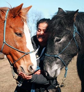 Anu stands between two horses that are facing each other. The horse on the left side of the picture is a reddish color with a white stripe down its nose and the right side horse is black with a gray nose. Anu is smiling and feeding the left horse something.
