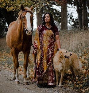 Anu stands on a dirt road with a reddish horse with a white stripe on his nose on her right. On her left is a golden retriever looking off to the side.