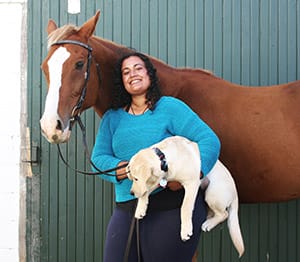 Anu stands in front of a green barn or shed door with Achates standing behind her. Anu is smiling and holding a young yellow lab puppy in her arms.