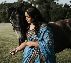 Anu's side is to the camera, and she is standing next to a dark colored horse that is looking toward her. They are outside is a grassy space with trees visible in the background.