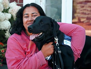 Anu sits on the front steps of a brick house. She is wearing a pink shirt and holding a black lab that's standing next to her. Bowman is wearing a blue Future Leader Dog vest.