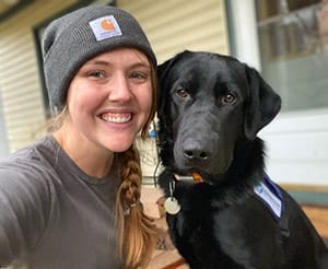 Ashley is smiling while taking a selfie with an adolescent black lab in a blue Future Leader Dog vest.