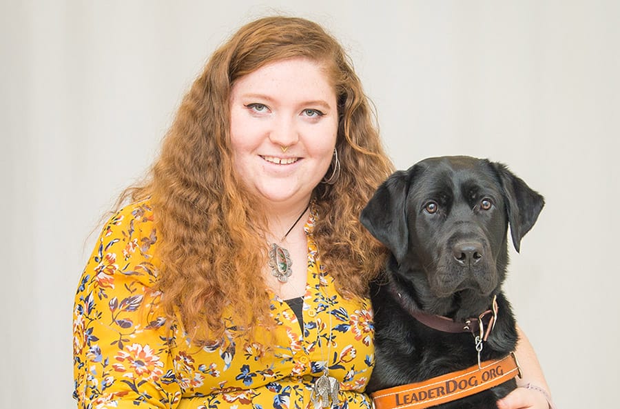 Cerr is seated next to black lab Iris. She is smiling at the camera and wearing a floral yellow top. Iris is in her Leader Dog harness. Behind them is a light gray photo backdrop.