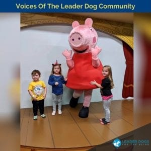 An adult in a pink pig costume is in the middle of a small stage. The pig’s hands are in the air and one foot is kicked off the ground. There are three children around the pig, two girls and one boy, who are looking at the camera and smiling.