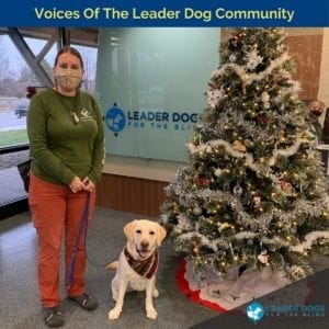 A woman wearing a green, long-sleeved LDB t-shirts, rust colored slacks and a patterned mask on her face is holding the leash of a yellow Labrador retriever in her left hand. The dog is wearing a red and green patterned holiday bandanna. They are standing in the lobby of the LDB canine center next to a Christmas tree with white garland and white lights.
