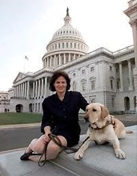 A woman with black hair just above her shoulders sitting on a ledge in front of the U.S. Capitol building holding a leash resting across her lap which is attached to the collar of a yellow Labrador retriever lying on the ledge next to her.