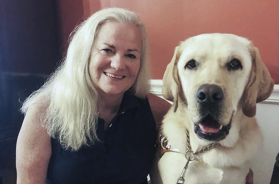 Moira sits smiling at the camera. Her long hair is down and she is wearing a black sleeveless top. Seated next to her is a yellow lab with its mouth slightly open as though smiling.