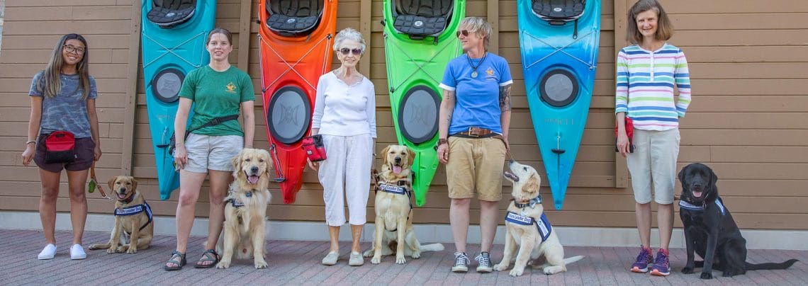 Five female puppy raisers with their dogs stand in front of a group of colorful kayaks.