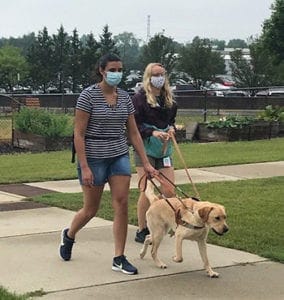 A young woman wearing shorts and a t-shirt walks with a yellow lab in Leader Dog harness. Next to the woman is another woman holding the dog's leash while they walk on a sidewalk surrounded by grass. Both women are wearing masks.