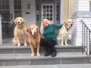 Moira sits on stone front steps in front of a residence. She's smiling and wearing a green sweater and black pants. On her left are two adult golden retrievers and on the right is a yellow lab.