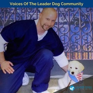 A man with a shaved head and goatee wearing a blue shirt, the same color pants with a white long sleeve under the top shirt is kneeling with his left hand on the back of a young yellow Labrador retriever puppy wearing a blue Future Leader Dog bandana. At the top of the photo is text reading Voices of the Leader Dog Community.