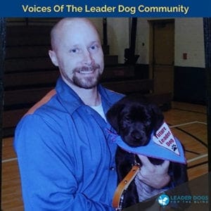 A man with a shaved haircut and goatee wearing a blue button down is holding a few week old black Labrador retriever puppy wearing a blue Future Leader Dog bandana.