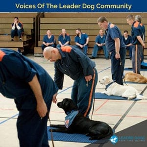 Inside a gymnasium are there are five men in a line each wearing a blue uniform with a orange stripe down the pants and across the back of the shoulders of the shirt. The man on the left is bending at the waist and his head is off camera. The man second to the left is bent over with his right hand in front of a black Labrador retriever wearing a blue Future Leader Dog bandana and lying down on a blue mat. The man in the middle is standing and has a light-colored yellow Labrador retriever lying down on his left wearing the same bandana on a blue mat. The man second from the right is standing with his left hand behind his back with a golden retriever wearing the same bandana and lying on a blue mat. The man all the way on the right has a black Labrador retriever wearing the bandana and lying on a blue mat on his left. There are six more men sitting in the bleachers in the background.