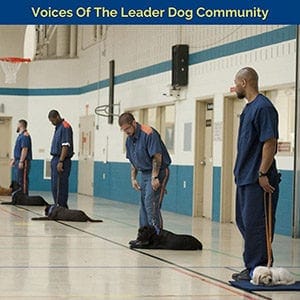 Line of four men in correctional facility outfits stand next to four dogs that are lying down next to them in a gym-like setting