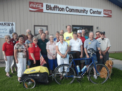 Mark stands with a group of people outside of a building with a banner on it that reads Bluffton Community Center. Mark's bicycle with its trailer is in front of them
