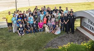 A group of 25 teenagers, male and female, some are holding canes in front of them, and some have canes on the ground next to them. There are also nine firefighters and 11 other adults standing with the teenagers in a large group. Next to them is a large sign with “Fire Station.”