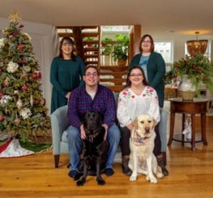 A group of three women and one man are in a living room with hardwood floors. They are all smiling and looking forward. There is a Christmas tree next to them. Two of the woman are standing behind a couch, a man and a woman are seated on the couch. The man has a black Labrador retriever seated in front of him, the woman on the couch has a yellow Labrador retriever seating in front of her.