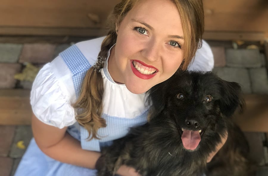 Ashley is wearing a Dorothy costume from the Wizard of Oz and smiling while she holds her pyrenean sheepdog, Gryf