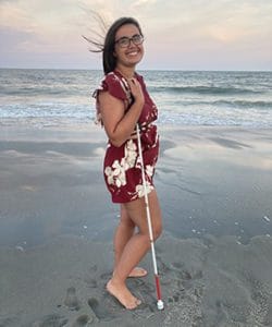 Brooke stands smiling on a beach with a white cane in her hand