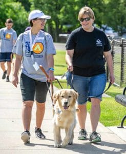 Brooke walks on a sidewalk with a golden retriever in harness. She is smiling and looking at the woman next to her, who is holding the leash